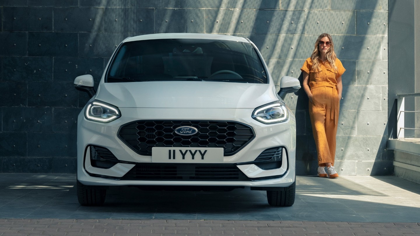 Ford Fiesta couleur blanche. Vue de face, stationnant devant un bâtiment moderne. Une femme se tient à côté.