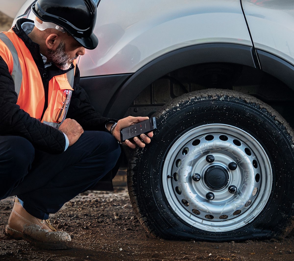 Un ouvrier portant un casque de chantier et un gilet de sécurité est agenouillé à côté d'un véhicule dont le pneu est crevé et tient un téléphone portable à la main.