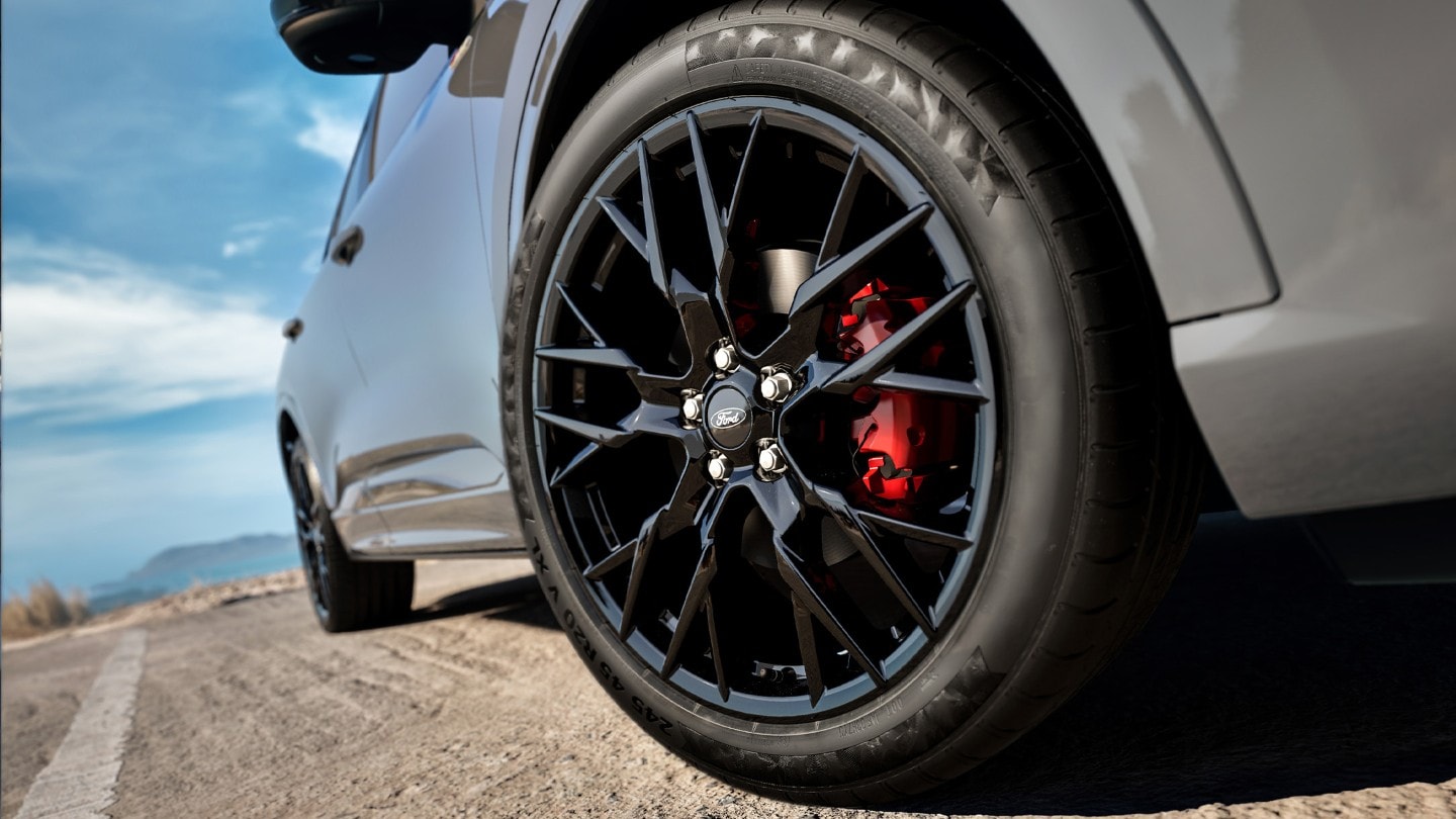 Roue de voiture grise sur asphalte, avec jante Ford noire et étrier de frein rouge, sous un ciel bleu.
