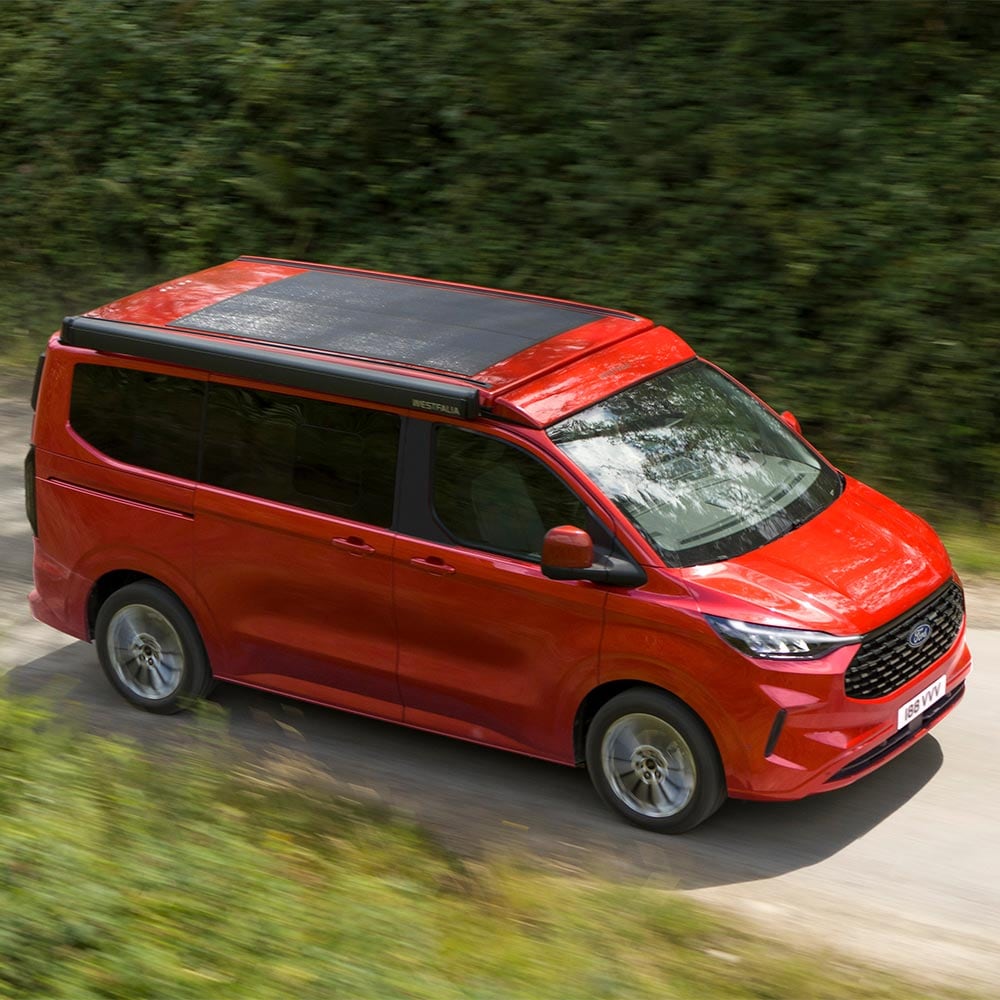 Ford Nugget rouge avec panneau solaire sur le toit circulant sur la route.