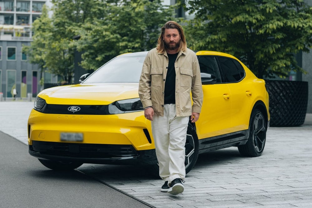 Un homme aux longs cheveux marche avec style devant une Ford Capri jaune