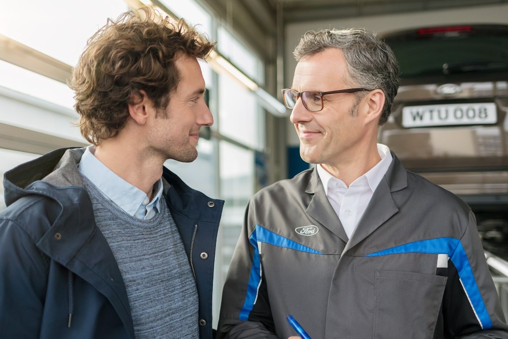 À l'atelier, un spécialiste du service Ford avec une tablette parle à un conducteur souriant dans la voiture.