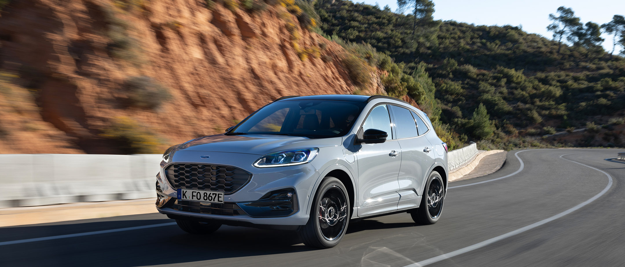 Ford Kuga couleur grise, vue de face aux trois quarts, roulant sur une route de campagne devant la pente d’une montagne