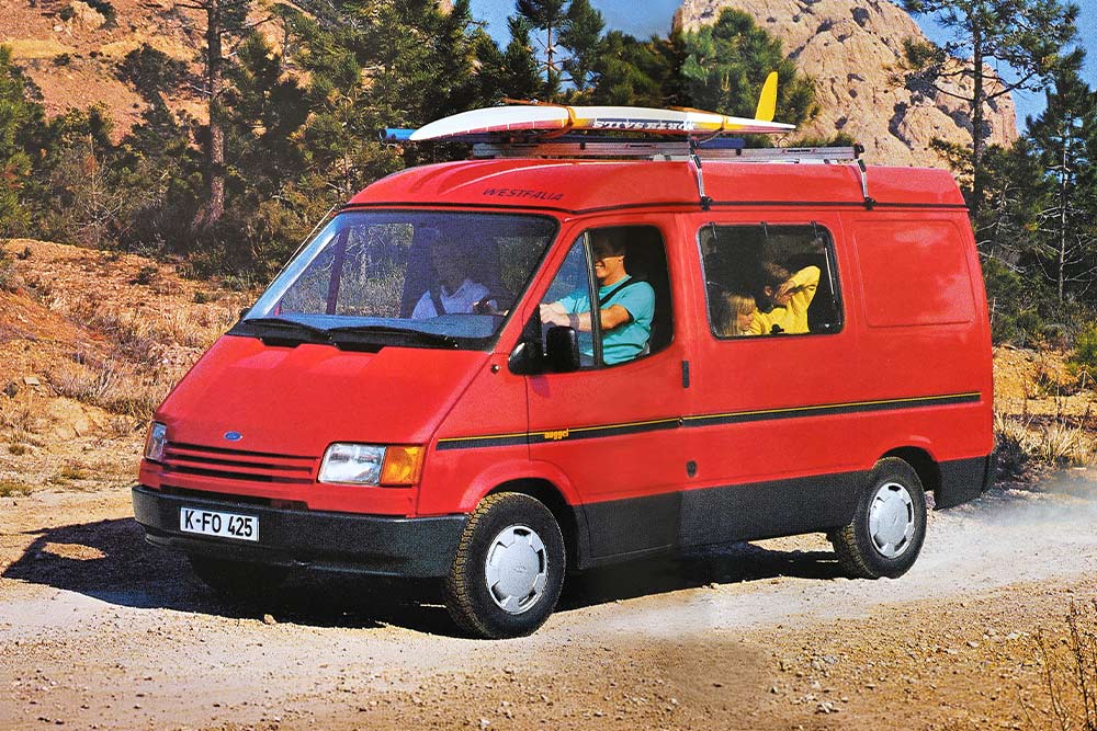 Un Ford Nugget de 1993 avec galerie et planche de surf sur une piste poussiéreuse en montagne.