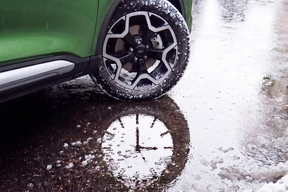 La roue avant d’un Ford Kuga vert se trouve à côté d’une flaque d’eau, à la surface de laquelle se reflète un cadran de montre.