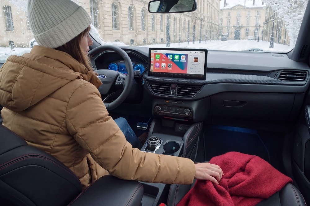 Une femme portant un manteau d’hiver et un bonnet est assise dans une Ford par une journée enneigée, tenant une couverture rouge.