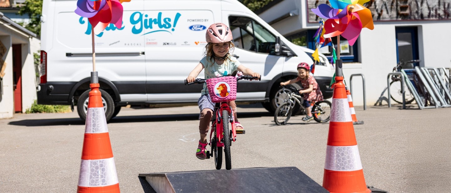 Une petite fille fait du vélo sur un parcours d'obstacles. En arrière-plan, une Ford E-Transit