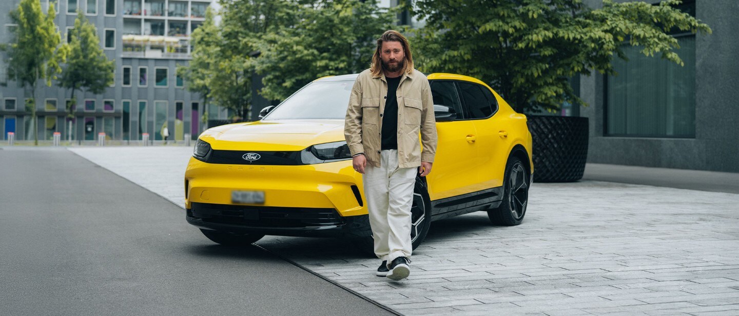 Un homme aux longs cheveux marche avec style devant une Ford Capri jaune