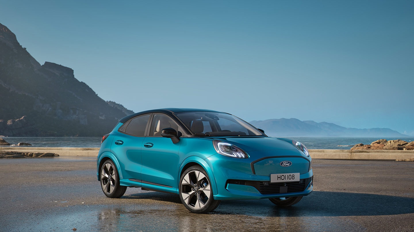 Vue de face d'un Ford Puma Gen-E bleu, stationné près d'un plan d'eau, avec des montagnes en arrière-plan.