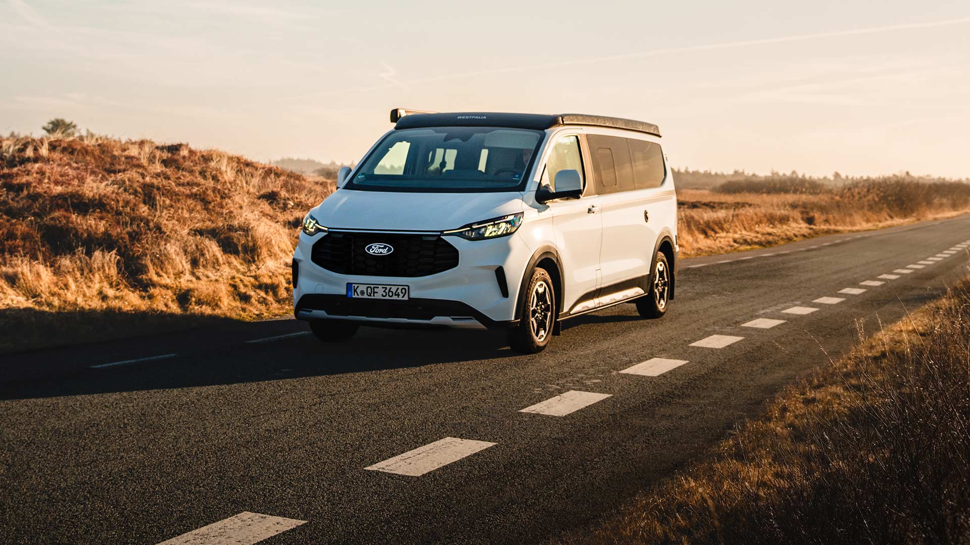 Vue de face du Ford Nugget édition anniversaire alors qu’il circule sur une route de campagne goudronnée, dans un paysage de dunes, sous le soleil.