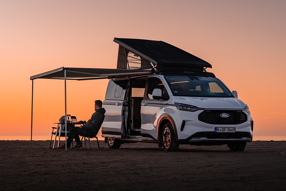 Ford Nugget édition anniversaire avec toit relevé et auvent déployé, dans un vaste paysage au crépuscule. Une personne est assise sur une chaise de camping sous l’auvent.