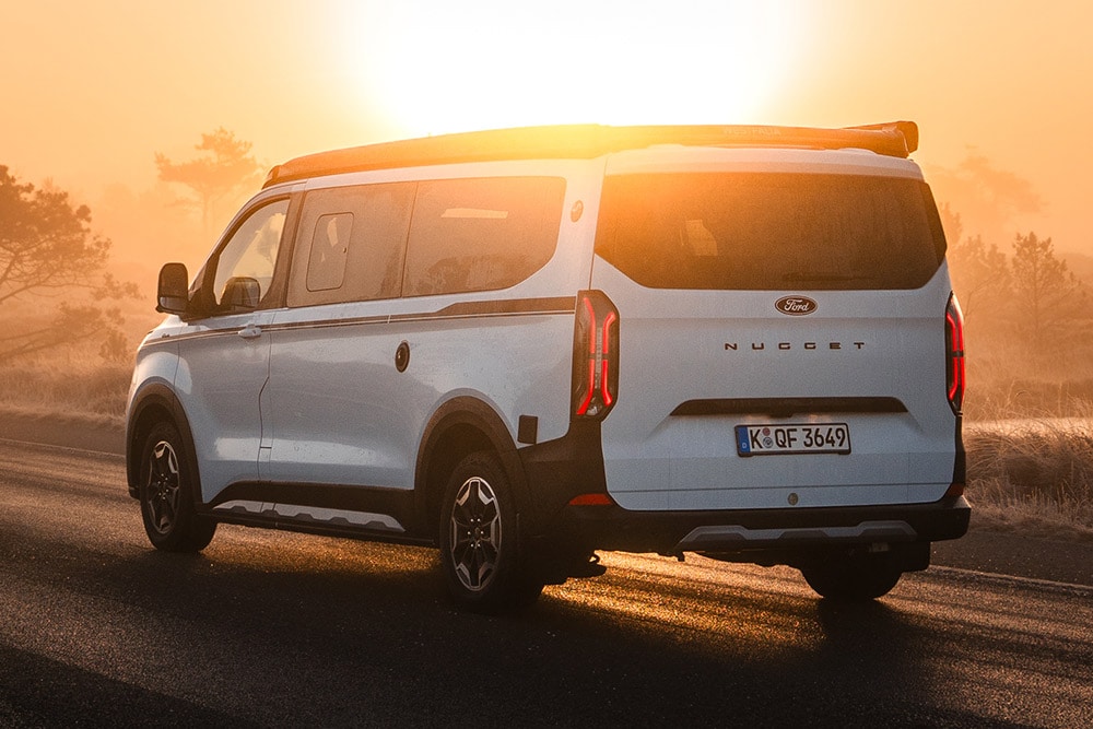 Vue arrière du Ford Nugget édition anniversaire sur une route goudronnée, dans un paysage brumeux au lever du soleil.