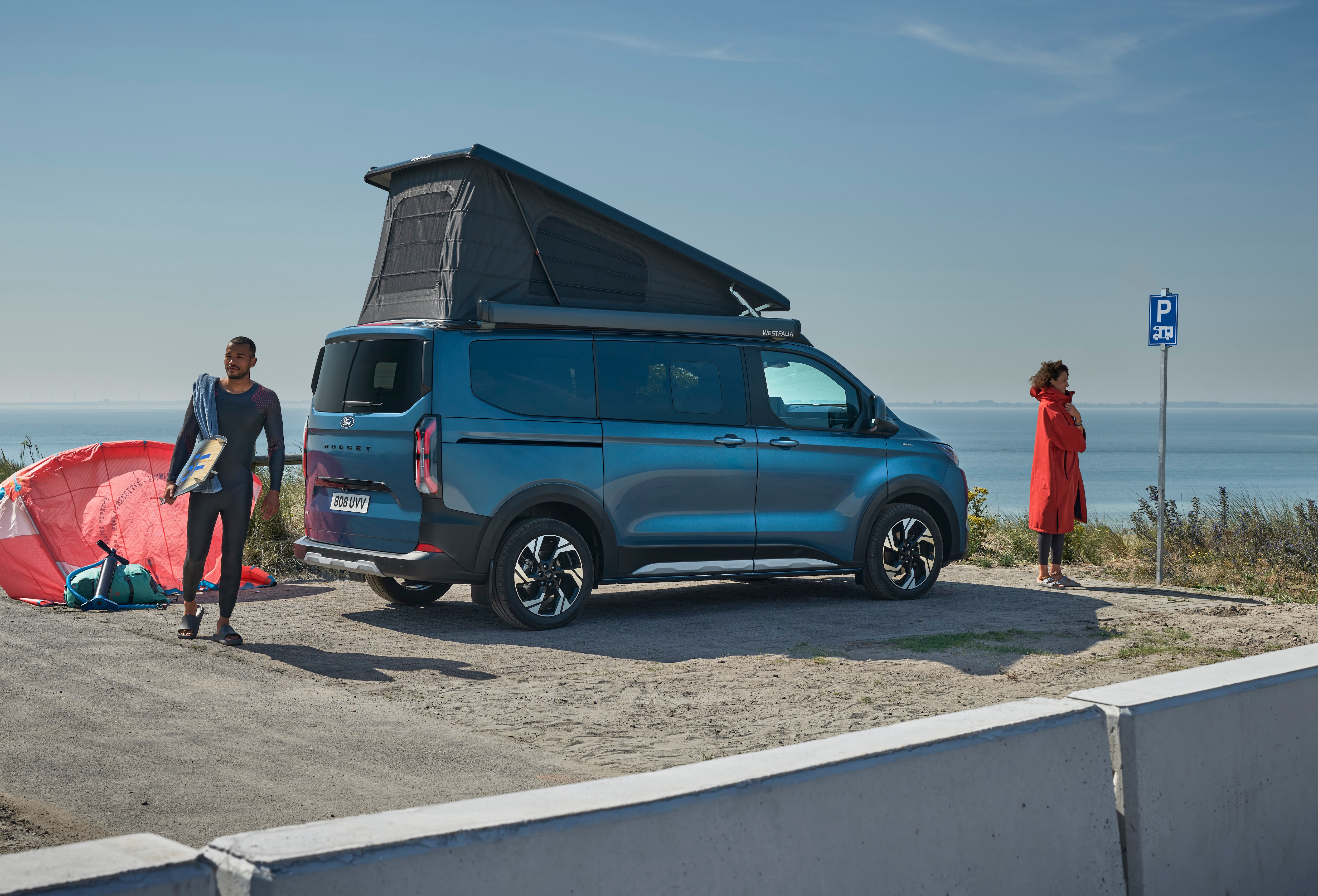 Un Ford Nugget bleu foncé avec toit relevé est garé au bord de la mer, alors que deux personnes préparent leur équipement de kitesurf.