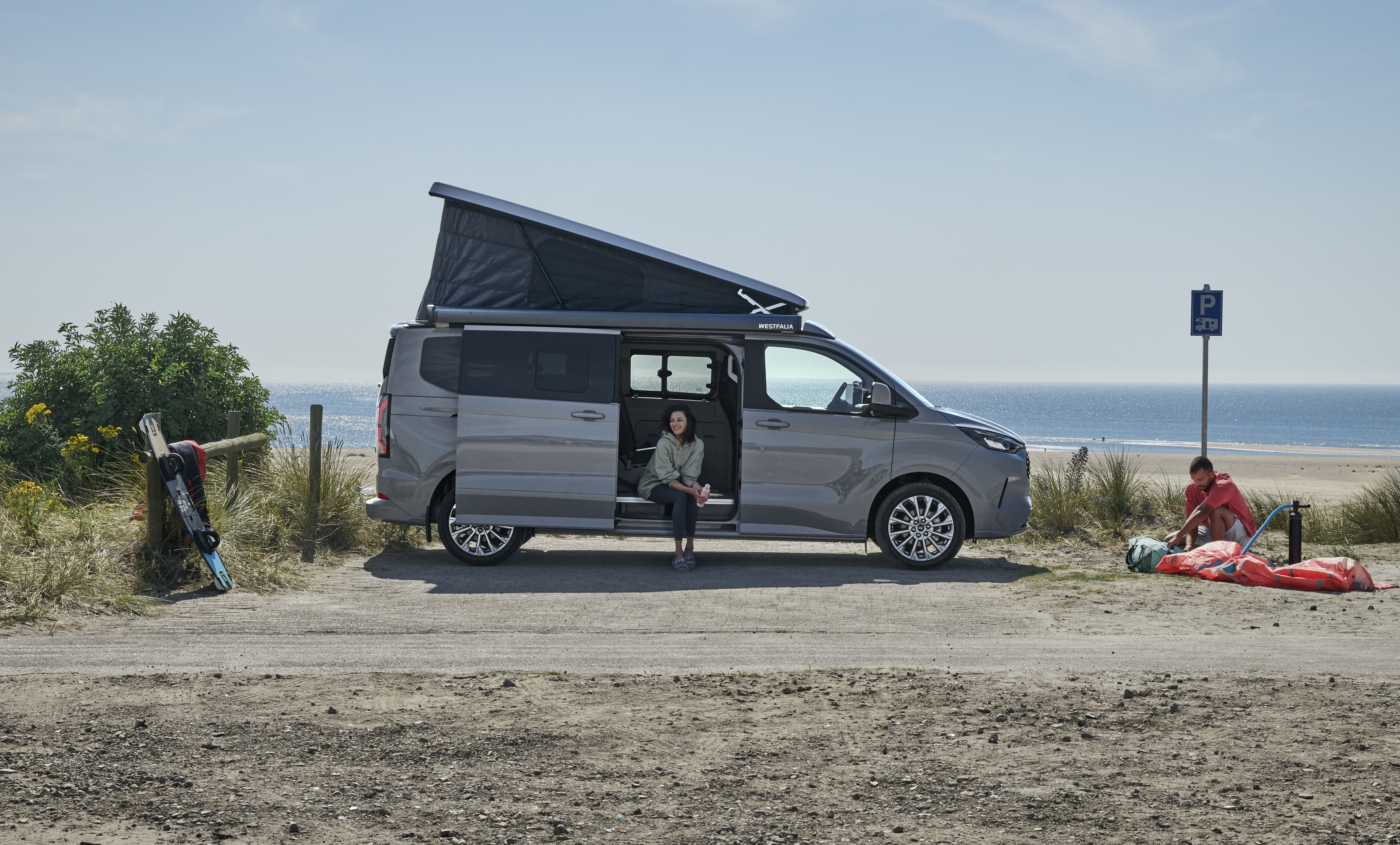 Un Ford Nugget gris avec toit relevé est garé à la plage; une personne est assise dans l’encadrement de la porte coulissante ouverte.