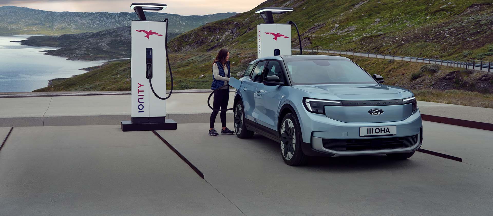 Une femme branche un Ford Explorer bleu, entièrement électrique, à une borne de recharge publique à l'aide d'un câble. La voiture est garée près de montagnes et d'eau.