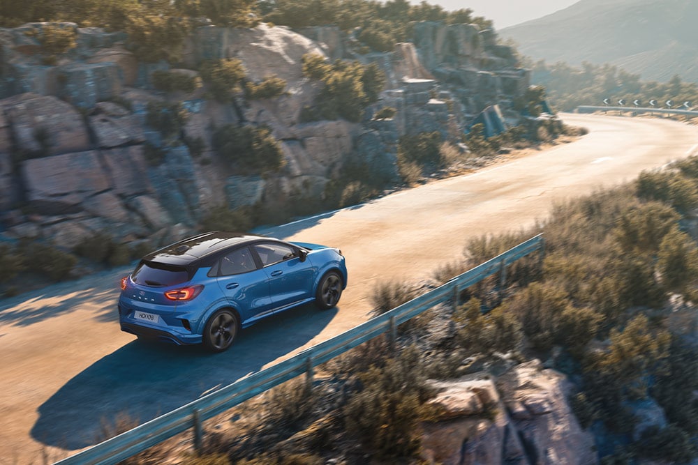 Un Puma Gen-E bleu roule le long d'une route de montagne sinueuse.