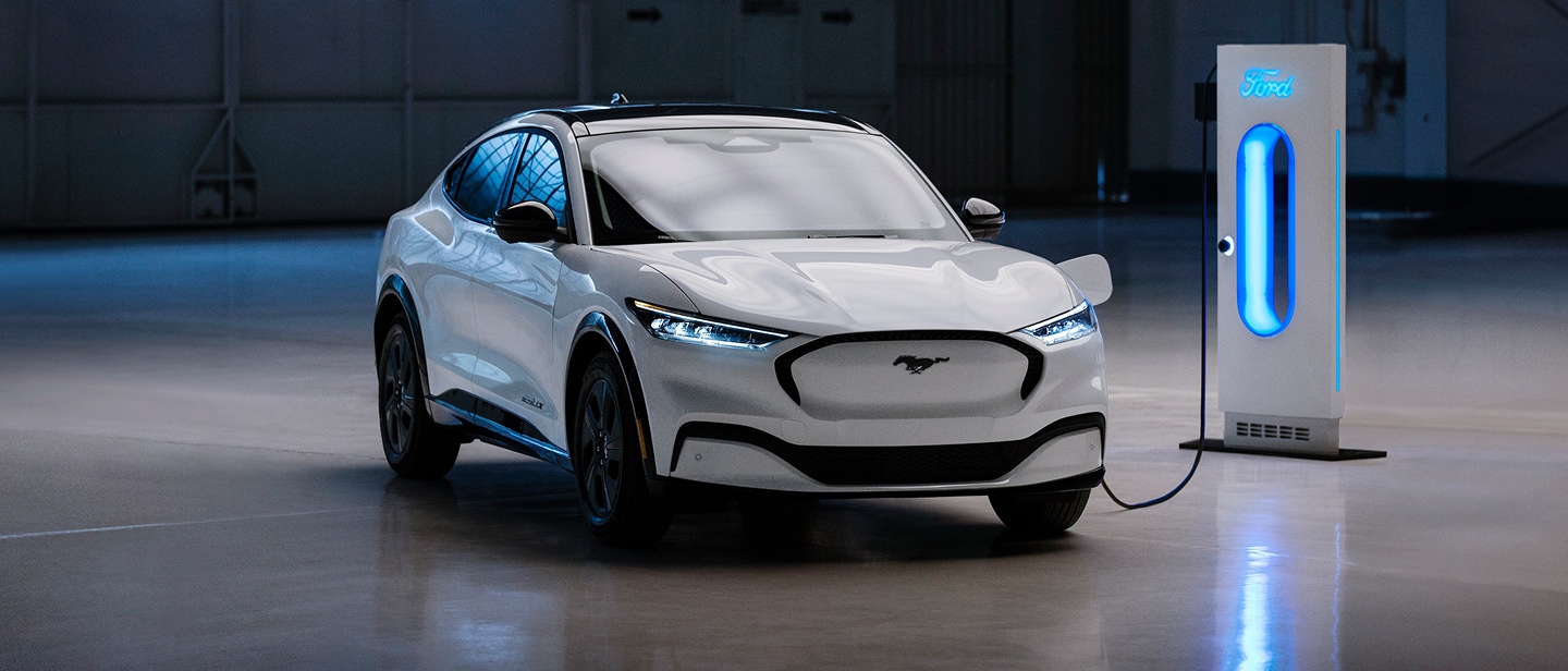 Une Ford Mustang Mach-E blanche est garée dans un garage et est connectée à une station de recharge éclairée en bleu.