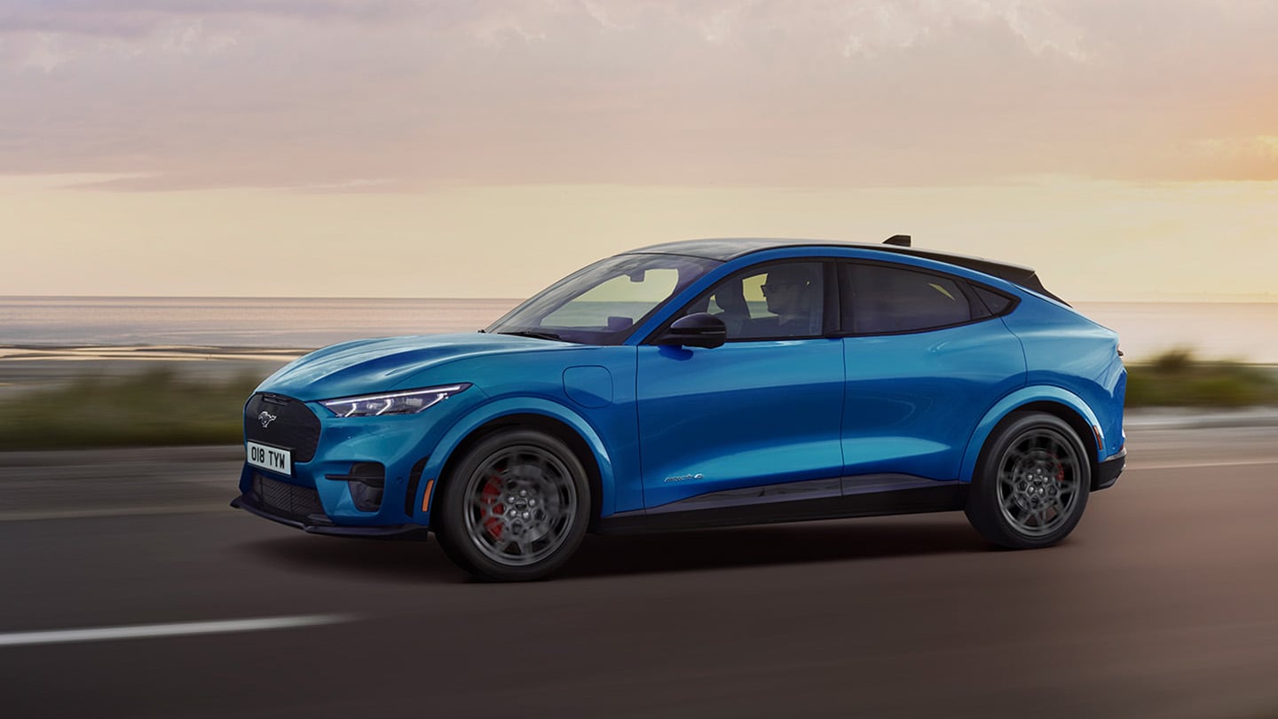 Vue latérale d’une Ford Mustang Mach-E électrique de couleur bleue roulant le long d’une route côtière au crépuscule.