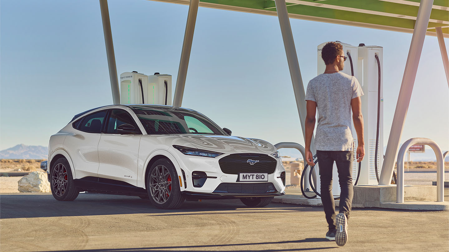 Une Ford Mustang Mach-E de couleur blanche stationnée devant une borne de recharge publique en plein air. Un homme vêtu d’un t-shirt blanc se dirige vers la voiture.