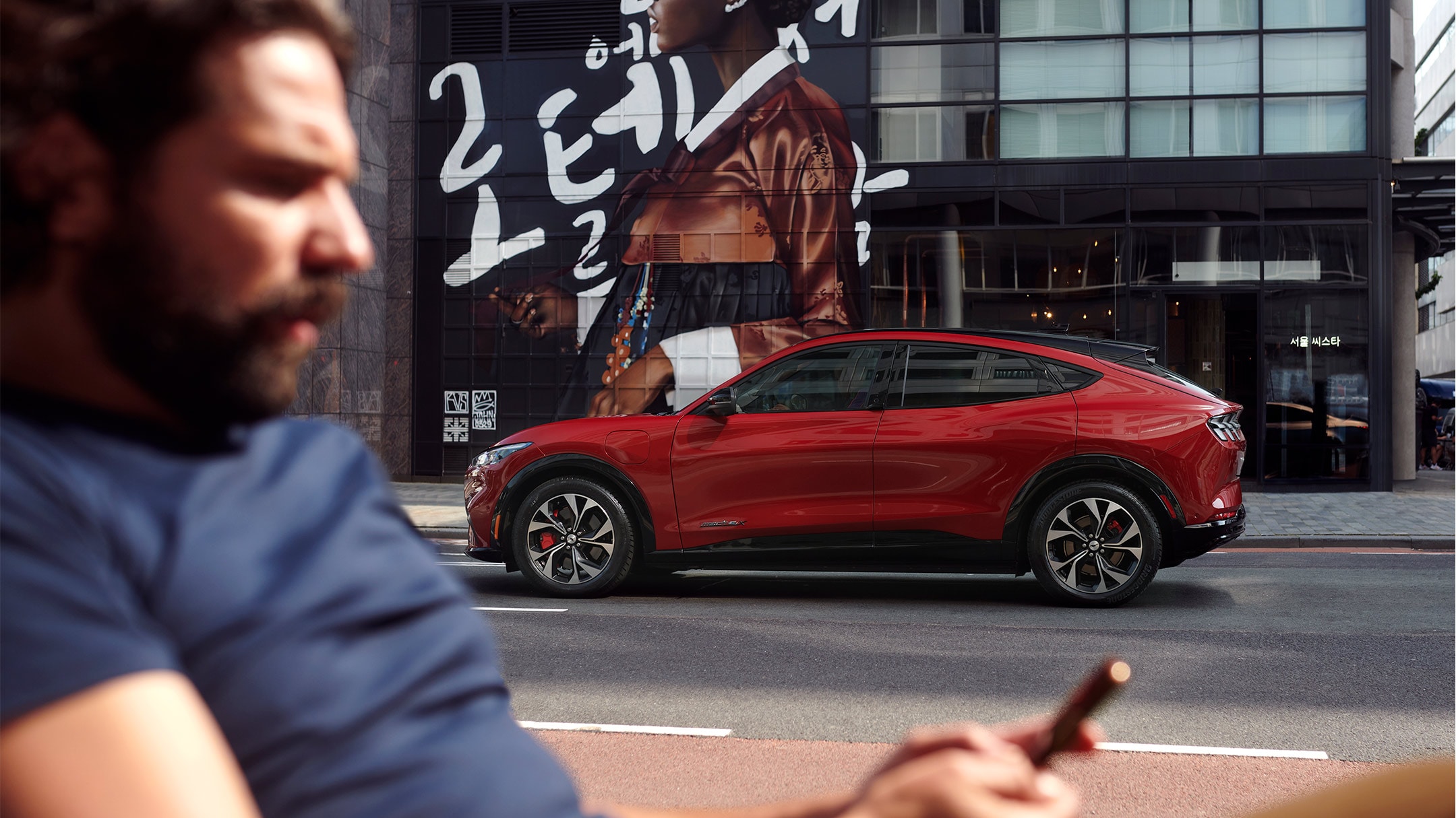 Ford Mustang Mach-E couleur rouge. Vue latérale. Un homme au premier plan qui regarde son smartphone