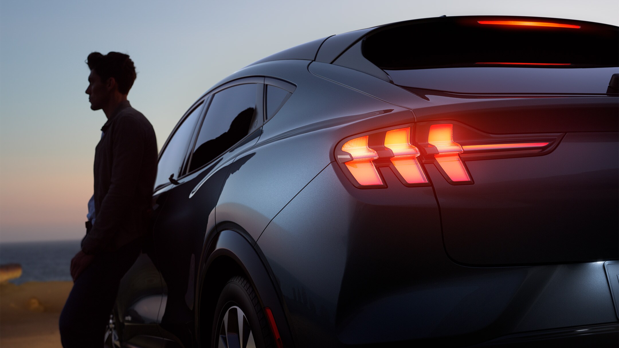 Ford Mustang Mach-E couleur grise. Homme appuyé contre son véhicule à la tombée de la nuit