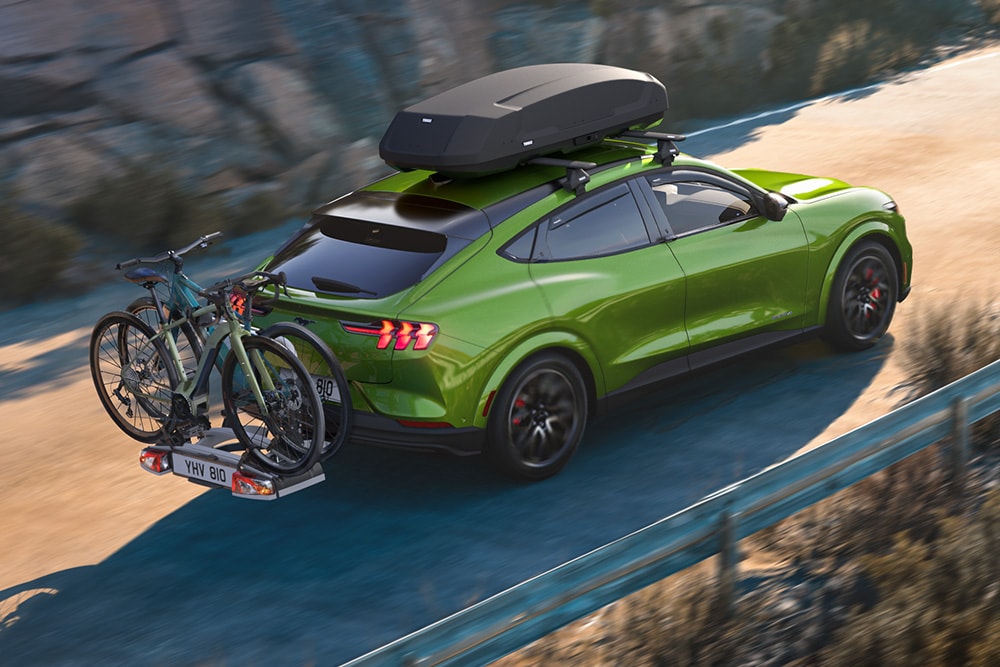 Ford Mustang Mach-E vert avec porte-vélos et coffre de toit sur une route sinueuse.
