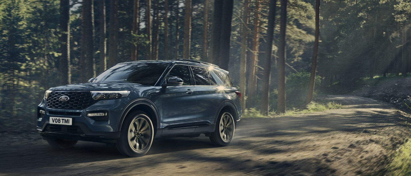 Ford Explorer Plug-In Hybrid bleu. Vue trois-quarts avant. Conduite sur une route de campagne avec une forêt en arrière-plan 