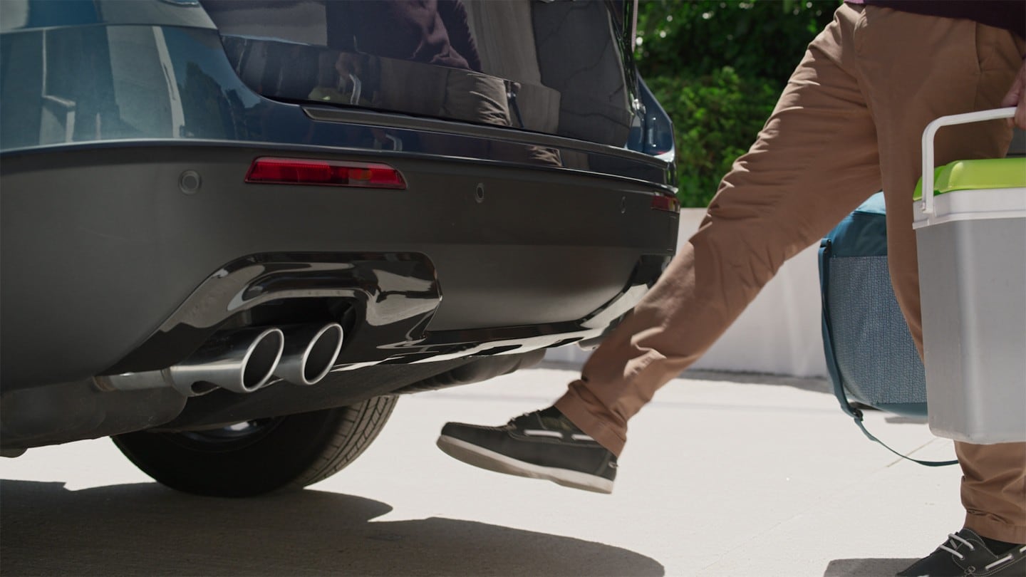 Ford Explorer Plug-In Hybrid bleu. Vue détaillée du hayon actionné par capteurs. Une personne avec les mains chargées déplace son pied sous le pare-chocs arrière.