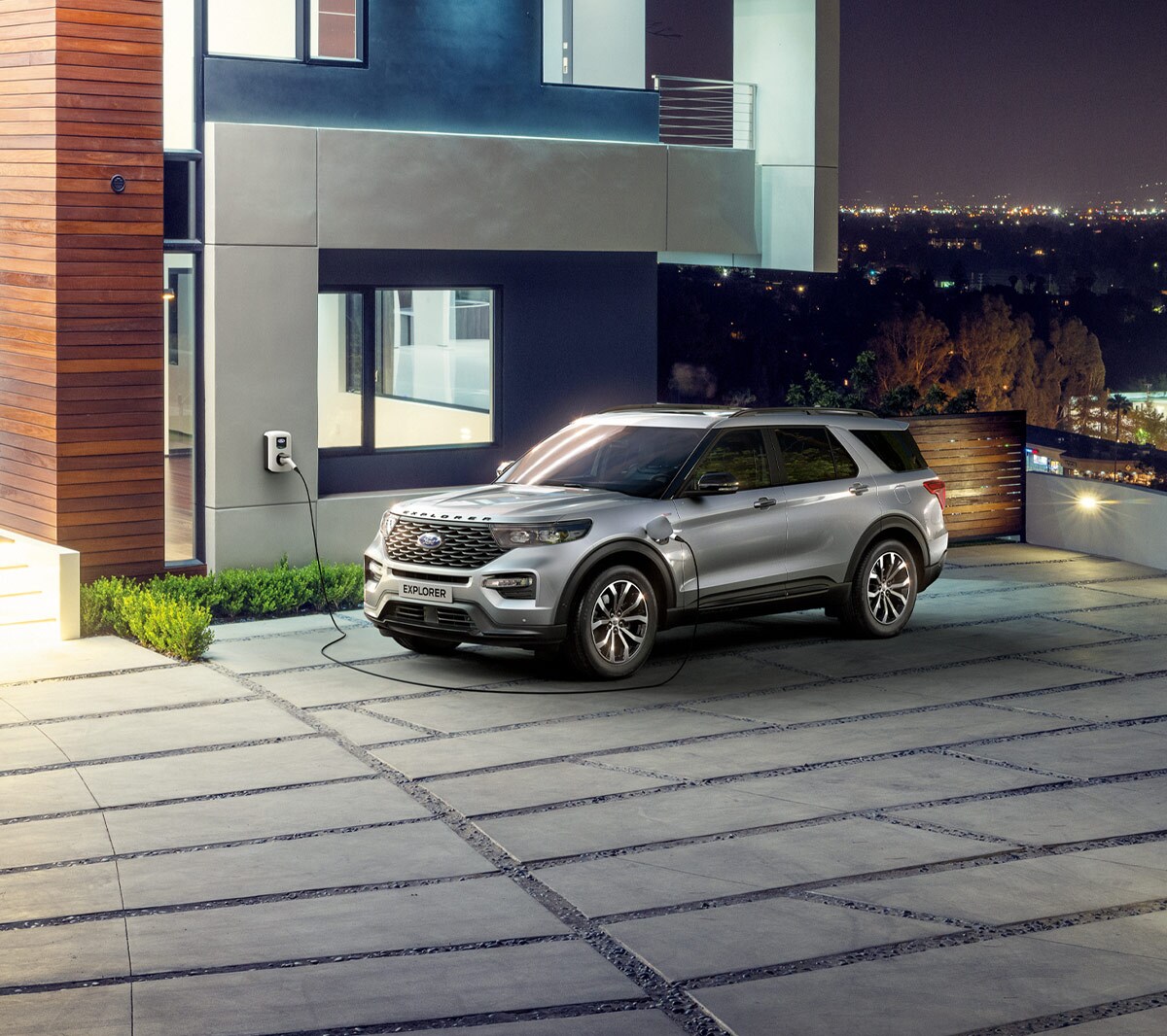 Ford Explorer Plug-In Hybrid de couleur argent. Vue trois-quarts avant. Stationnement devant une maison particulière. Chargement à une station de charge