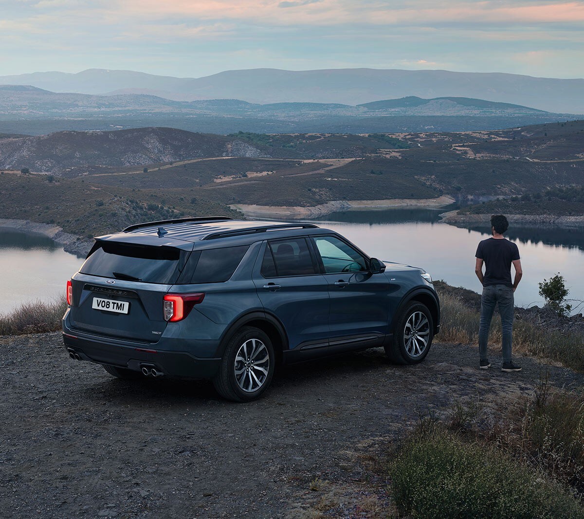 Ford Explorer Plu-In Hybrid bleu. Vue trois-quarts arrière. Stationnement dans une région collineuse, avec une personne à côté du véhicule admirant le paysage 