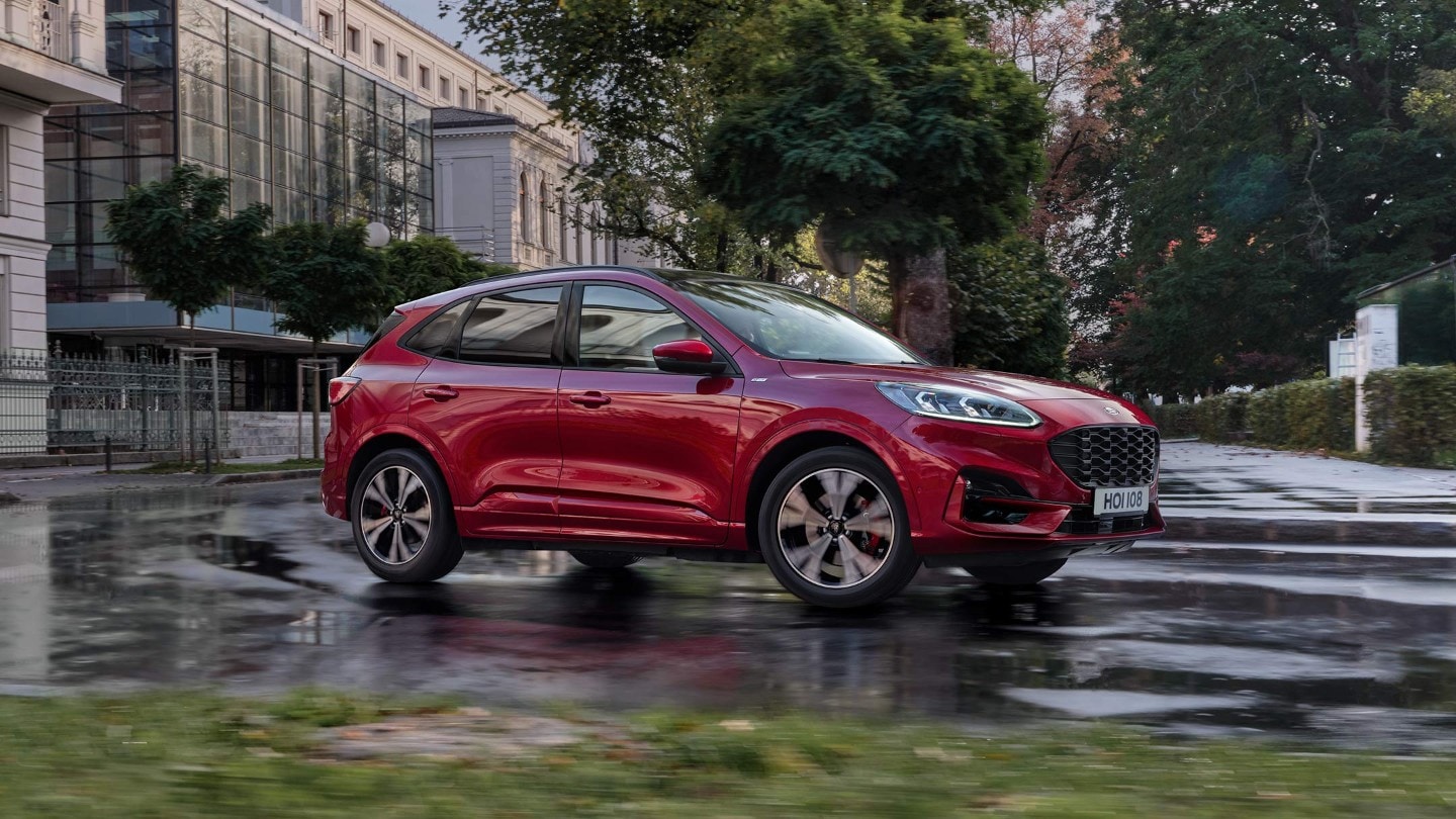 Ford Kuga rouge, vue de face aux trois quarts, en ville, roulant devant des bâtiments modernes et des arbres