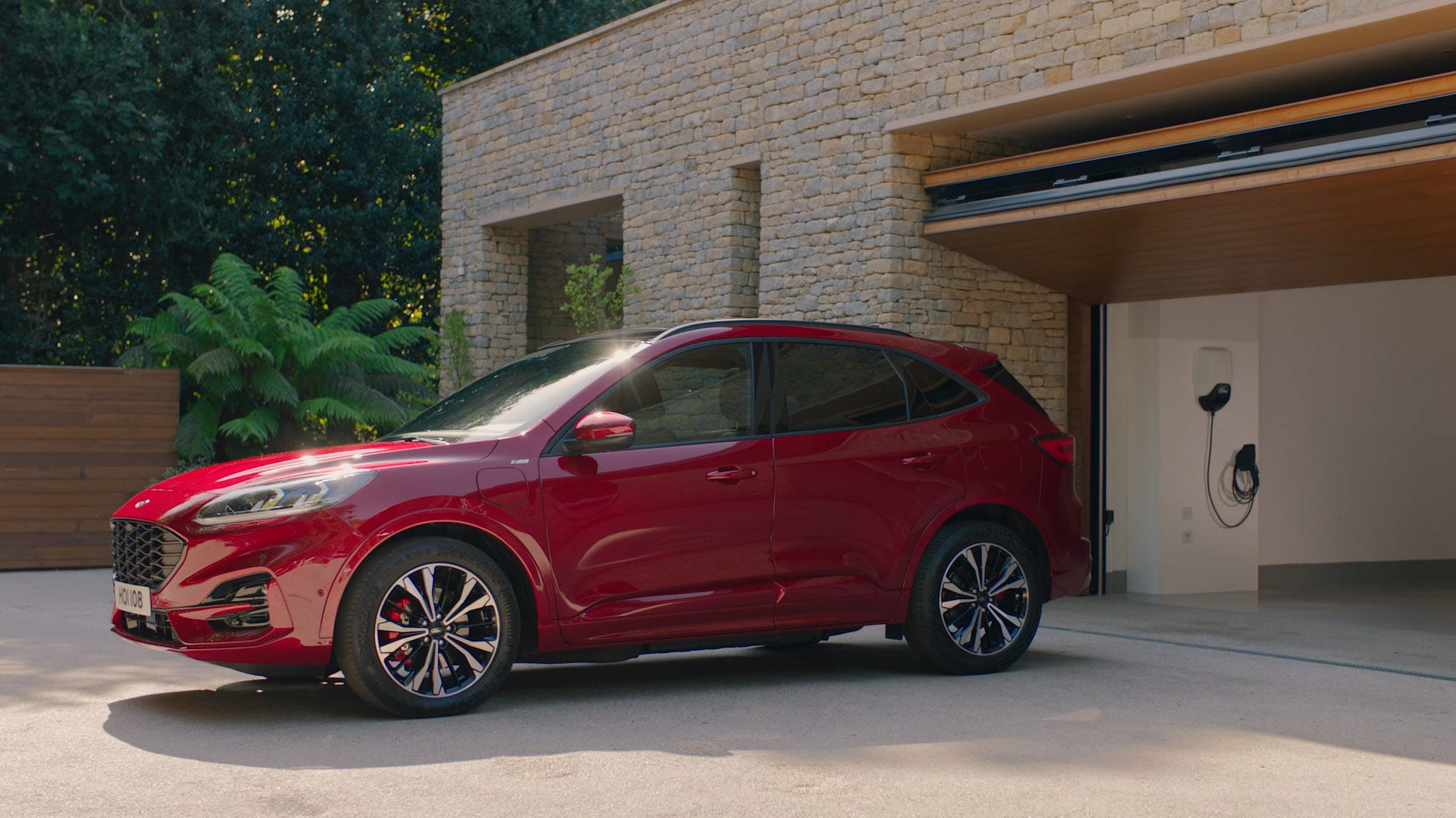 Ford Kuga Plug-in Hybrid (PHEV) couleur rouge. Vue latérale, devant un garage ouvert avec Wallbox