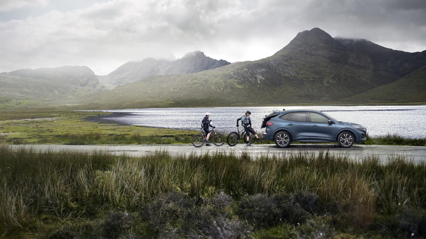 Ford Kuga couleur bleue. Vue latérale, garé devant un lac en montagne. Un cycliste de retour de sa balade ouvre le hayon arrière commandé par capteurs à l’aide d’un mouvement du pied