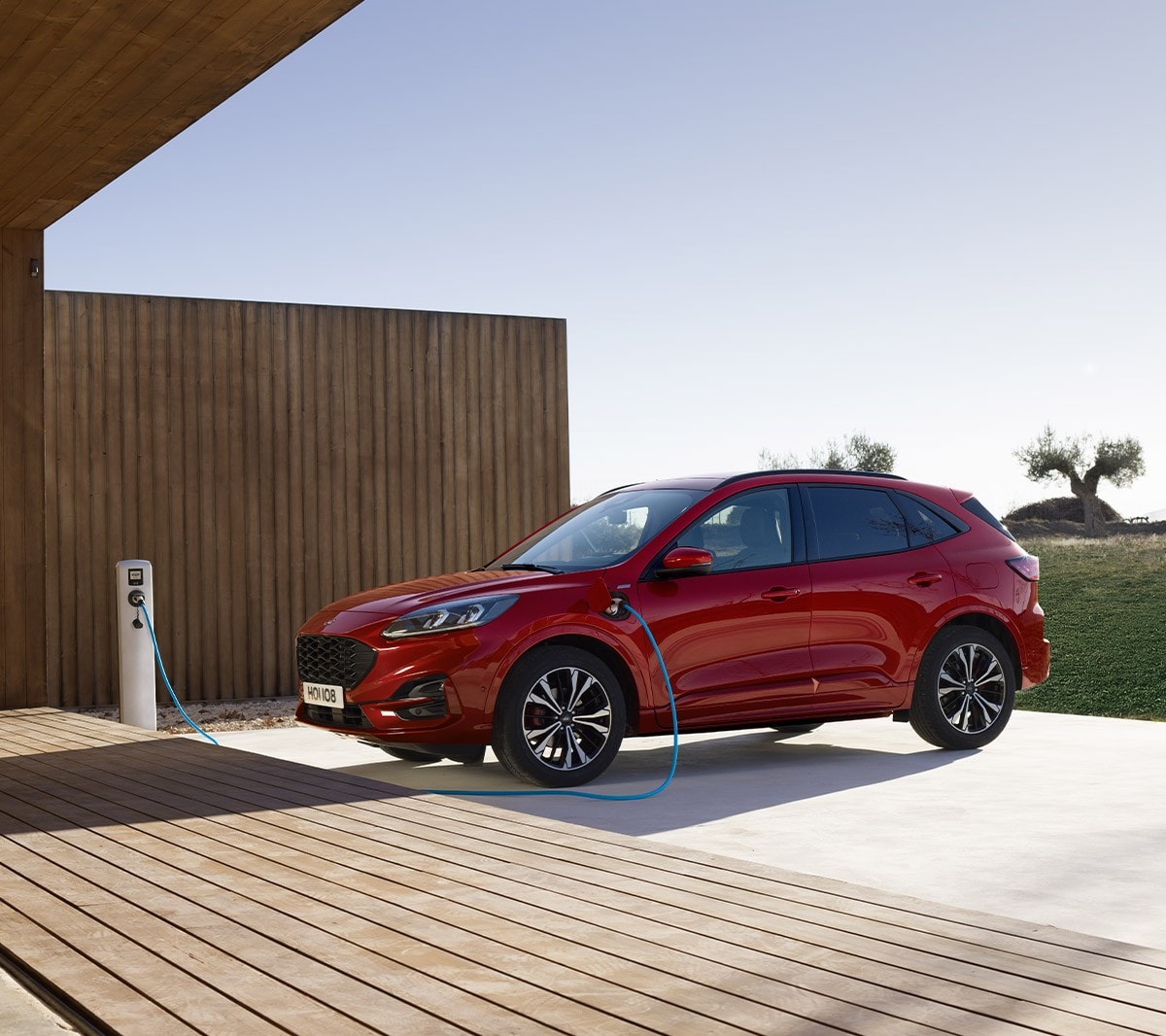 Ford Kuga couleur rouge. Vue latérale, à une station de recharge, devant un bâtiment moderne