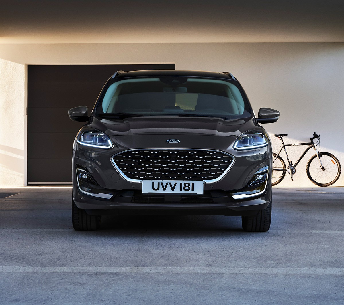 Ford Kuga couleur bleue. Vue de face, garé devant un garage sous l’avant-toit d’un bâtiment moderne.