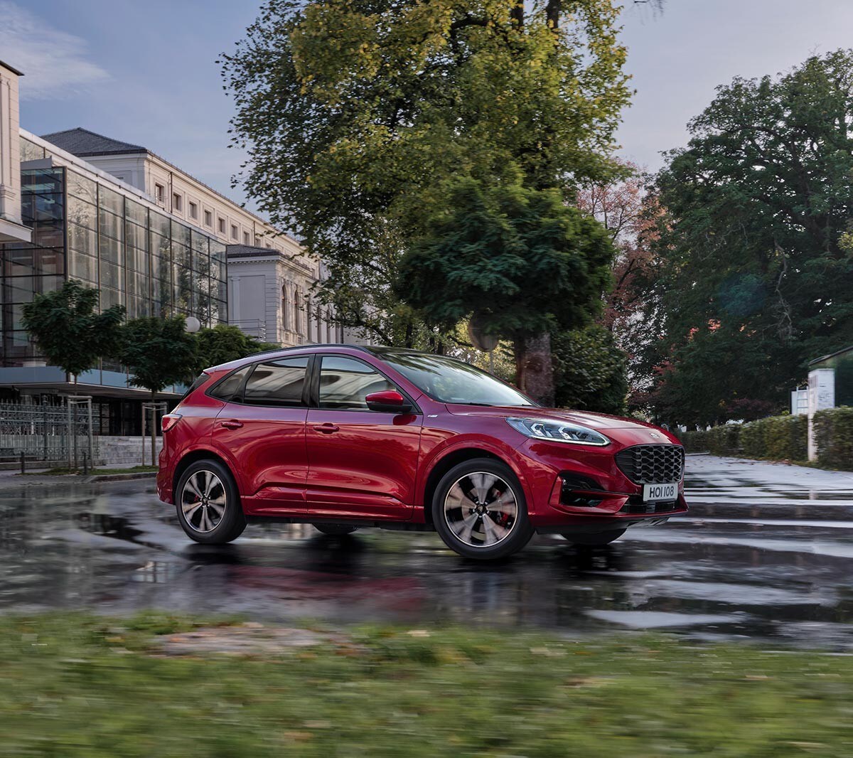 Ford Kuga couleur rouge. Vue de face aux trois quarts, roulant en ville devant un bâtiment moderne