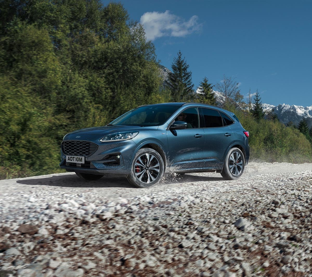 Ford Kuga couleur bleue. Vue de face aux trois quarts, roulant sur un chemin de gravier devant une forêt