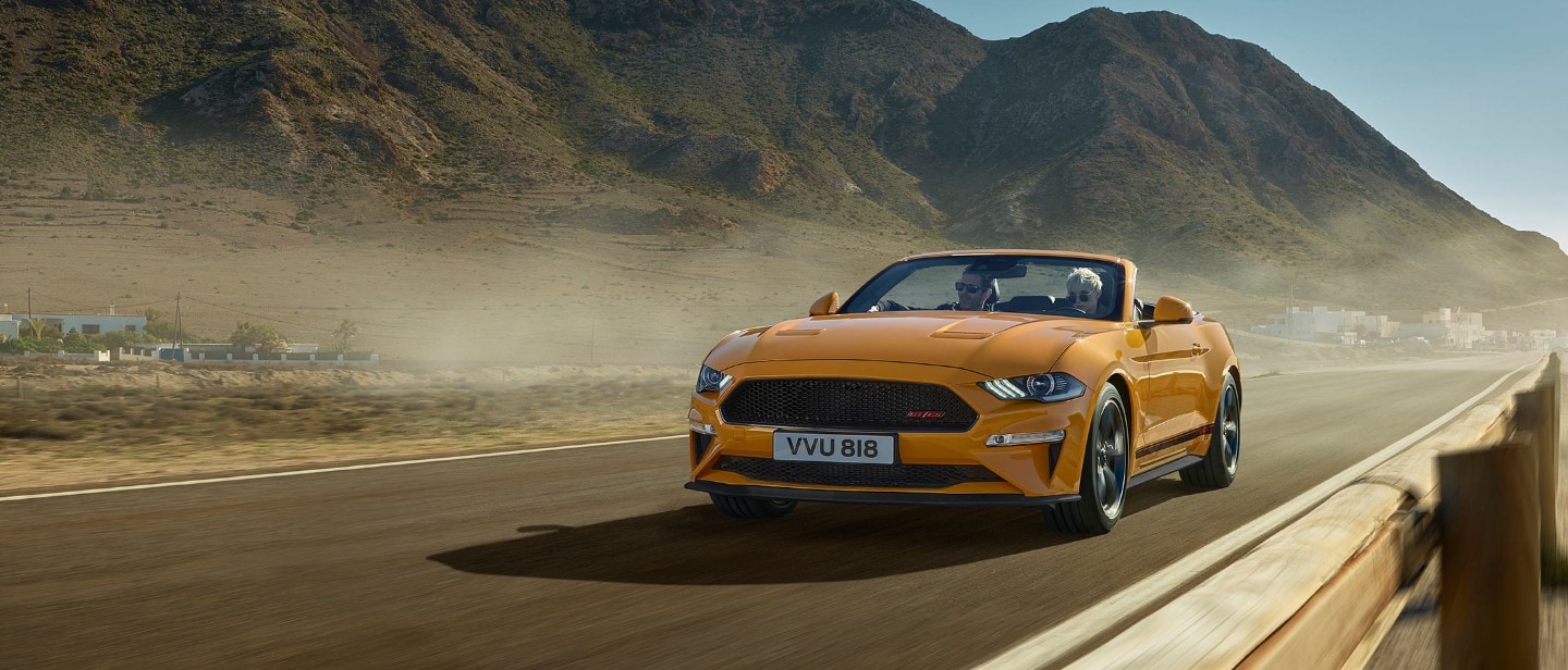 Ford Mustang California couleur orange. Vue de face aux trois quarts, roulant dans la nature avec des montagnes en arrière-plan.