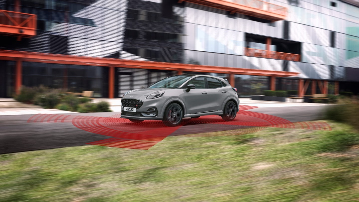 Ford Puma ST couleur grise. Vue de face aux trois quarts, roulant dans une rue avec un bâtiment moderne en arrière-plan et l’assistant de conduite visualisé