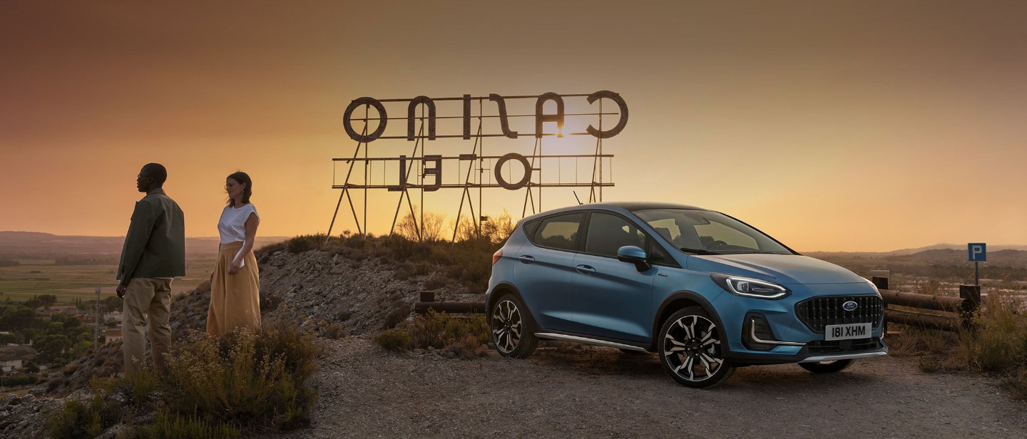 Ford Fiesta couleur bleue. Vue de face aux trois quarts, stationnant dans la nature. Deux personnes se tiennent à côté.