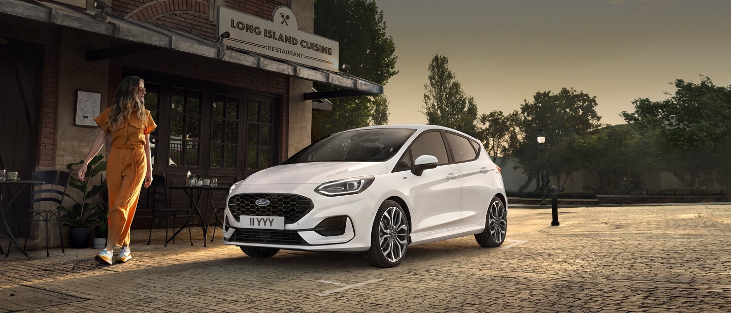 Ford Fiesta couleur blanche. Vue de face aux trois quarts, stationnant devant un restaurant avec une femme au premier plan.