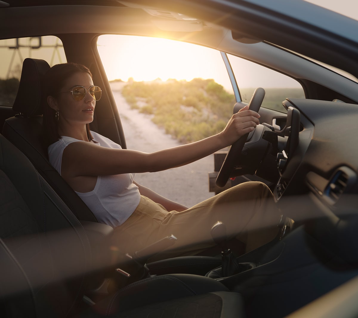Ford Fiesta. Vue intérieure. Une femme est assise sur le siège conducteur avec la porte ouverte de son côté. Un coucher de soleil à l’arrière-plan.