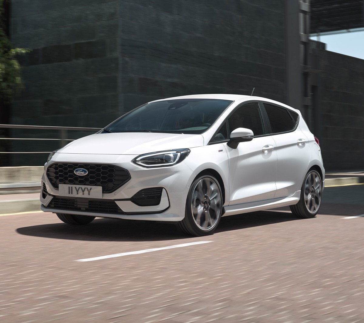 Ford Fiesta couleur blanche. Vue de face aux trois quarts, roulant en ville avec des bâtiments modernes à l’arrière-plan.