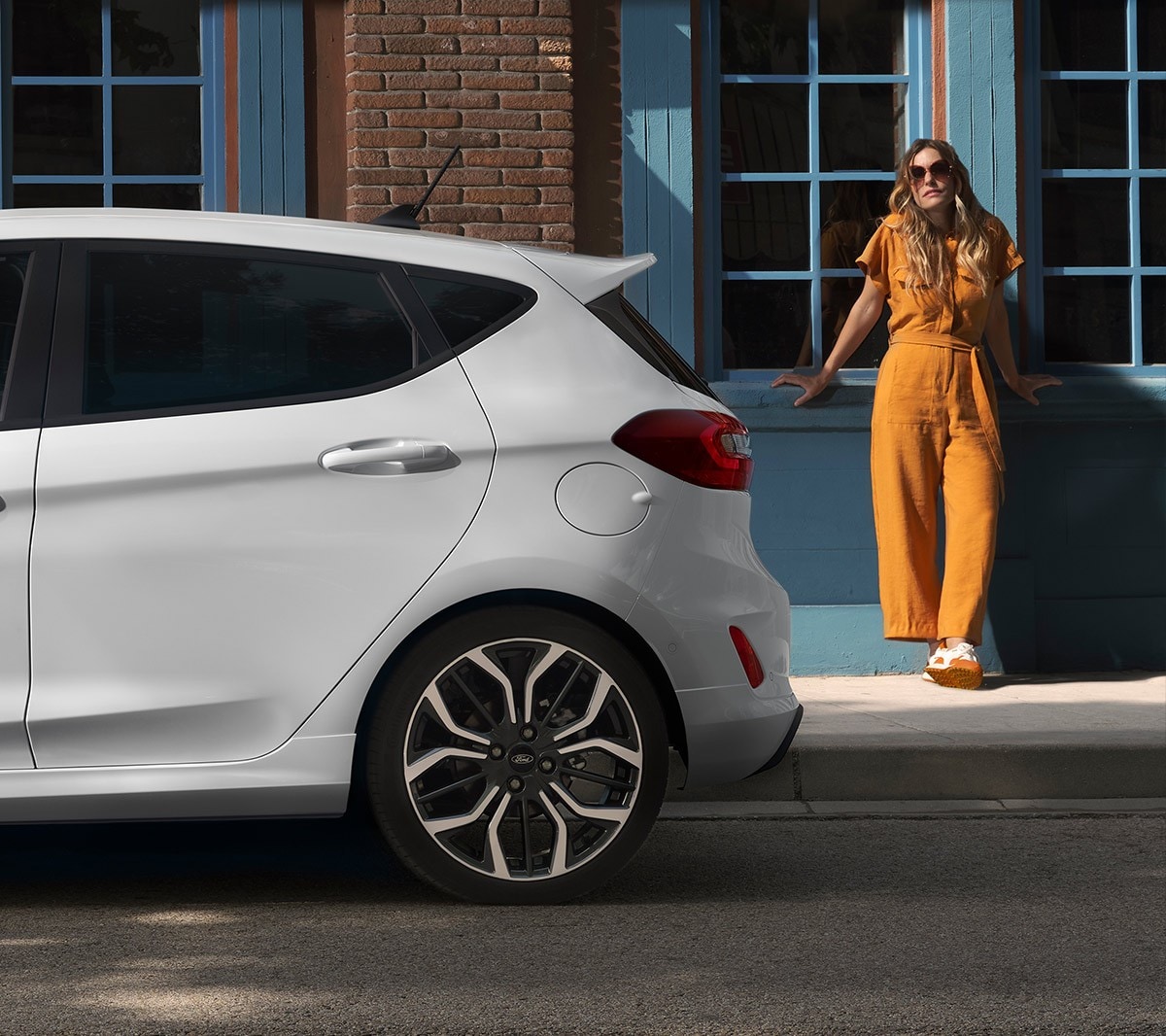 Ford Fiesta couleur blanche. Vue latérale, stationnant devant une maison avec une femme à l’arrière-plan.