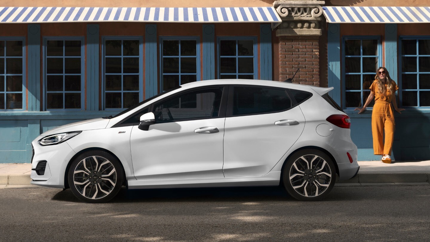 Ford Fiesta couleur blanche. Vue latérale, stationnant devant une maison avec une femme à l’arrière-plan.