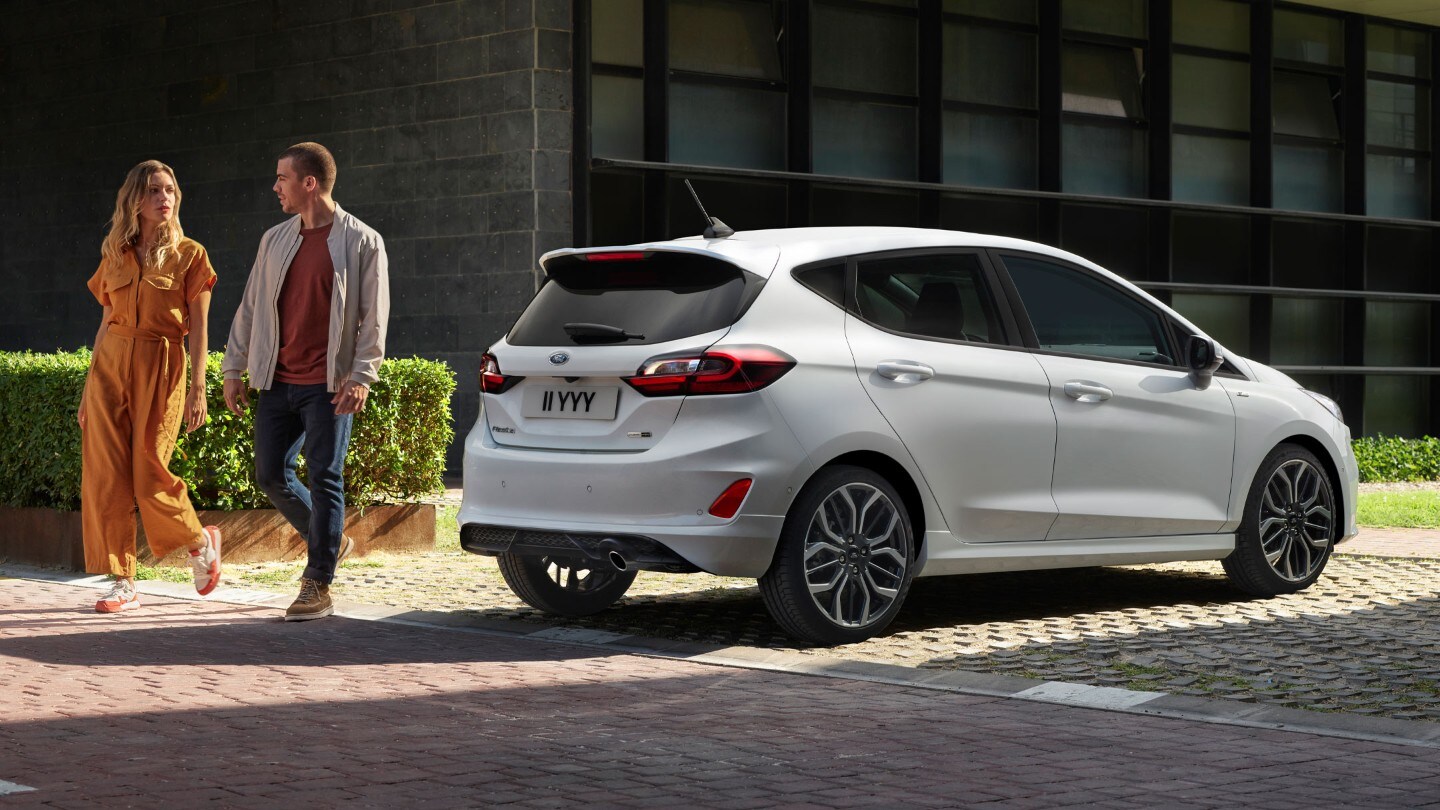 Ford Fiesta couleur blanche, vue arrière aux trois quarts, stationnant devant des bâtiments avec deux personnes à côté.