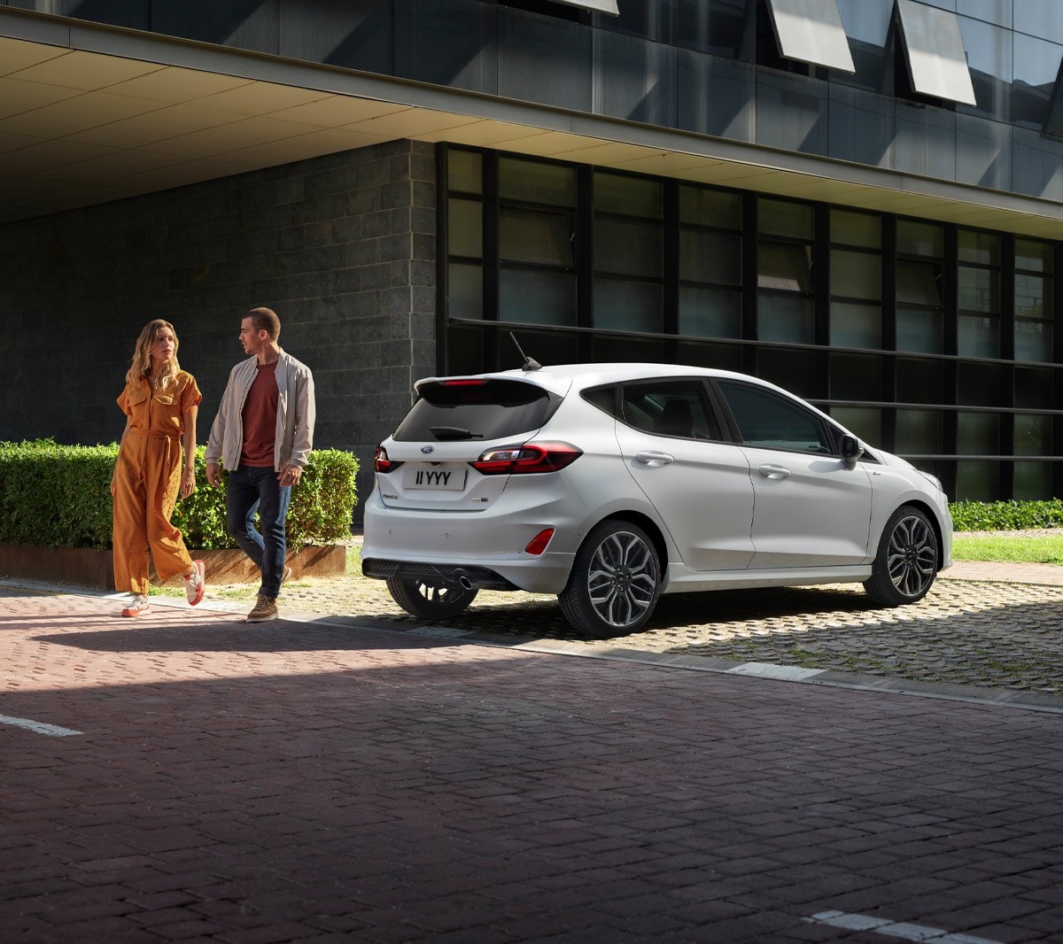 Ford Fiesta couleur blanche. Vue arrière aux trois quarts, stationnant devant des bâtiments avec deux personnes à côté.