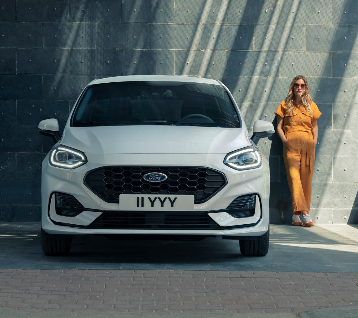 Ford Fiesta couleur blanche. Vue de face stationnant devant un bâtiment moderne, avec une femme à l’arrière-plan.