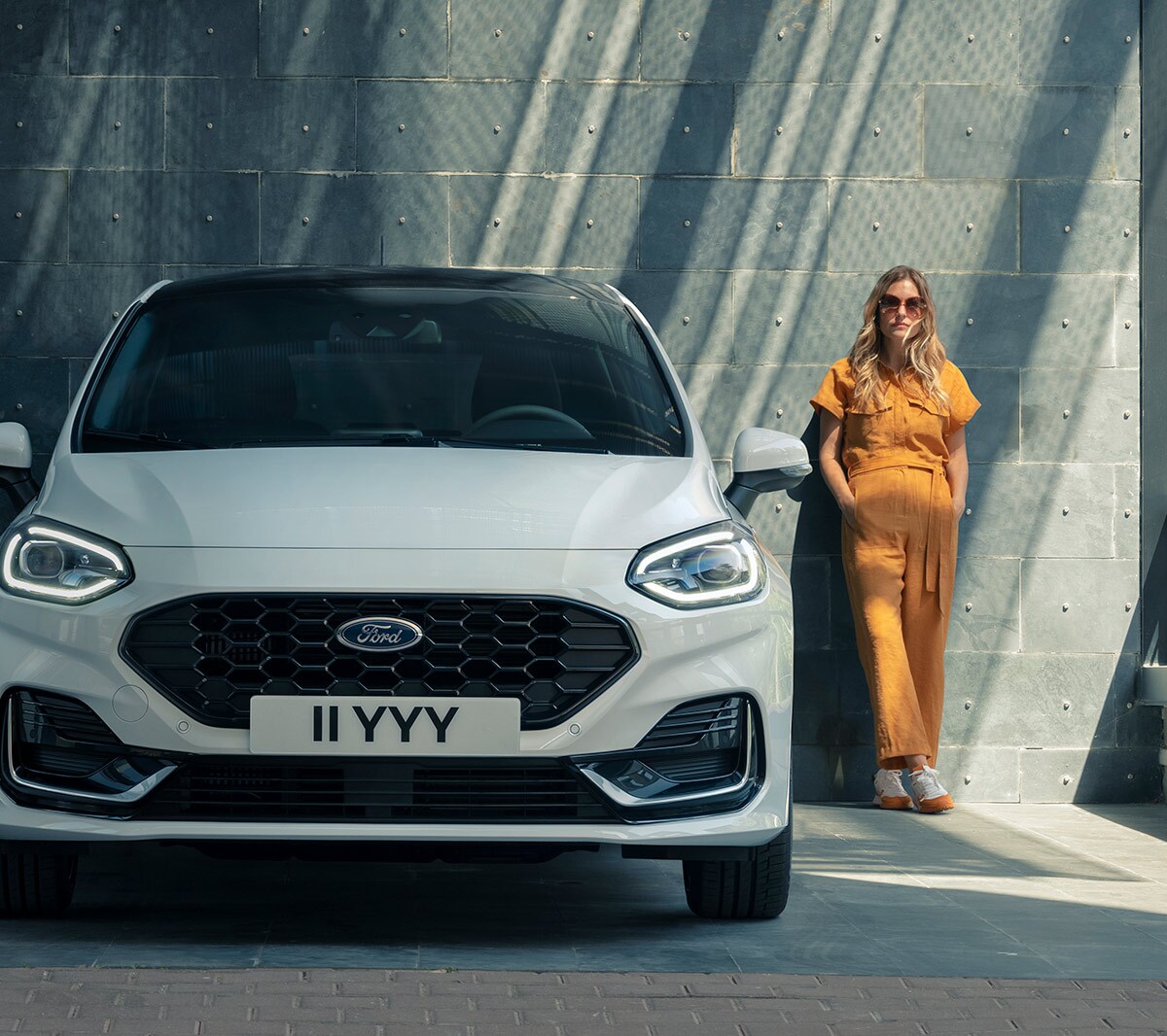 Ford Fiesta couleur blanche. Vue de face stationnant devant un bâtiment moderne, avec une femme à l’arrière-plan.