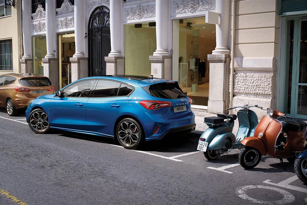 Une Ford Focus de couleur bleue se gare dans une place de parking en ville à l’aide du système d’aide au stationnement actif.